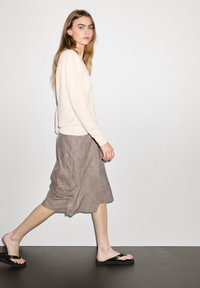 Maglione beige con polsini a coste, gonna grigia in suede con tasche e infradito nere, modella che cammina su uno sfondo neutro.