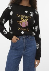 Svart stickad tröja med vita prickar och snöflingor, med guldfärgade paljetter som formar texten "GOD JUL" och en rosa presentgrafik.