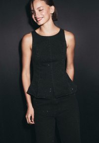 Czarny top typu peplum wykonany z fakturowanej tkaniny, z delikatnymi błyskotkami i dopasowanym krojem, noszony z pasującymi czarnymi spodniami.