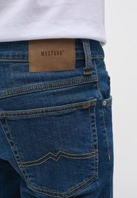Mėlyni džinsai su tvirtu tekstūru, rudi odiniai ženkleliai su "MUSTANG" prekės ženklu ir geltonos siūlės detalėmis.