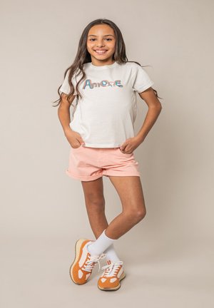 Fille debout souriante avec de longs cheveux bruns, portant un t-shirt blanc avec le texte « Amore », un short rose, des chaussettes blanches et des baskets orange.