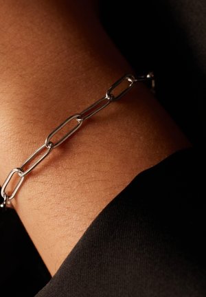 Zilveren kettingarmband met verlengde schakels, tentoongesteld op een gebruinde pols tegen een donkere achtergrond; gladde textuur en reflecterende afwerking.