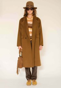 Long manteau en laine brun avec un col cranté, associé à un pull rayé, un pantalon en simili cuir marron et des baskets en daim jaune. Chapeau marron et lunettes de soleil.