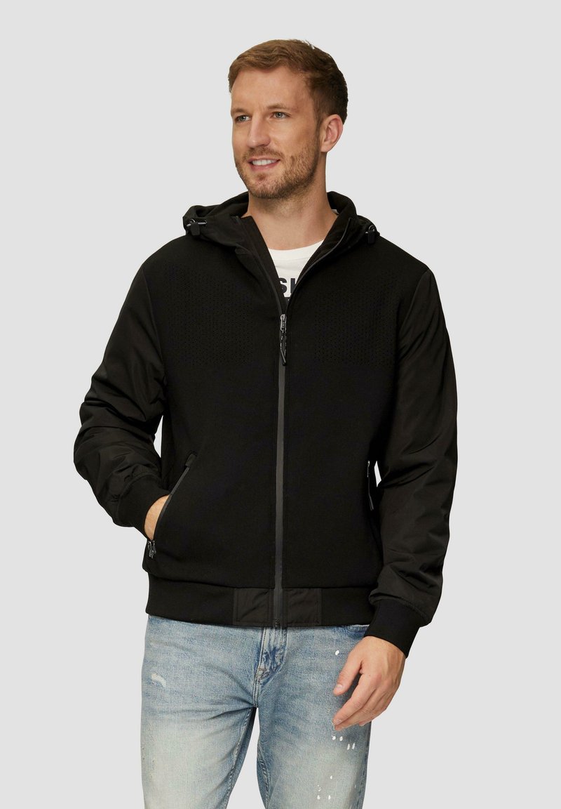 Veste noire zippée avec une surface texturée, capuche et deux poches avant, associée à un jean déchiré bleu clair.