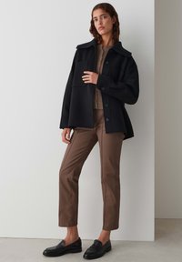Veste en laine mélangée noire oversize avec grand col et fermeture à boutons, associée à un pantalon droit marron et des mocassins noirs. Haut en tricot texturé en dessous.