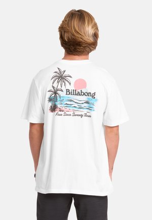 Person mit kurzen blonden Haaren, die ein weißes Billabong T-Shirt trägt, das auf der Rückseite einen Sonnenuntergang, Palmen, Meereswellen und Text zeigt.