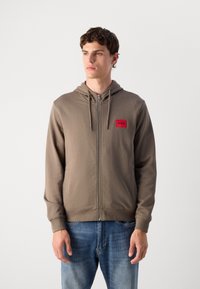 Chaqueta con capucha y cremallera en mezcla de algodón marrón con un parche de logo rojo en el lado izquierdo del pecho, que cuenta con bolsillos laterales y puños acanalados.