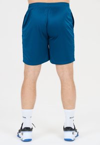Man som bär blå idrottsshorts, vita strumpor och svartvita sneakers, står vänd bort från kameran på en vit bakgrund.