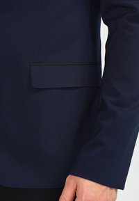 Mörkblå kavaj med ärm och ficklock med svart trim, bärs av en person, där armen och handen är delvis synliga.