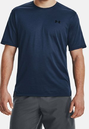 Mann trägt ein marineblaues, kurzärmeliges Sportshirt mit schwarzem Under Armour-Logo auf der Brust und graue Shorts, steht mit entspannten Armen.