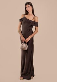 Braunes, bodenlanges Kleid mit schulterfreien Ärmeln und Spaghettiträgern. Glattes Material, kombiniert mit einer hellen Clutch-Tasche und Plateausandalen.