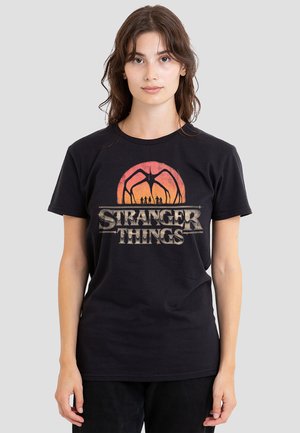 Young woman wearing a black Stranger Things t-shirt featuring a sunset and shadowy creature with four small figures underneath.