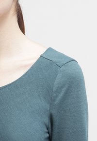 Haut texturé bleu sarcelle avec un col carré et des épaules ajustées, présentant un tissu lisse qui capte subtilement la lumière.