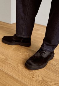 Scarpe nere realizzate in materiale testurizzato, con una superficie liscia e suola in gomma. Abbinate a pantaloni scuri a righe sottili su un pavimento in legno.