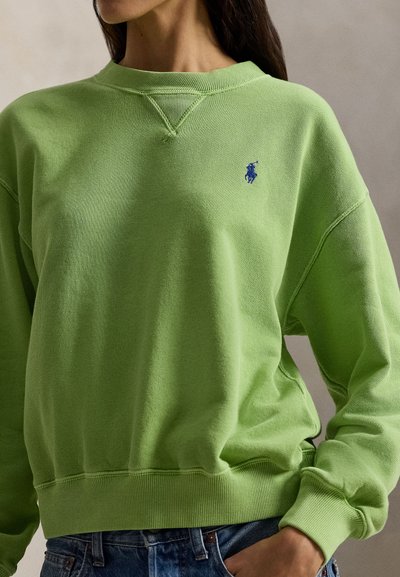 Kvinde iført en limegrøn sweatshirt med et lille blåt broderet logo på brystet, kombineret med blå denimjeans.