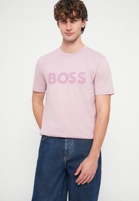 Gaiši rozā īsroku T-krekls ar tekstu "BOSS" tumšākā rozā tonī, kopā ar tumši zilām džinsa biksēm.