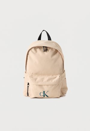 Μπεζ σακίδιο πλάτης Calvin Klein με μαύρη λαβή, εμπρόσθια τσέπη με φερμουάρ και λογότυπο "ck Calvin Klein" σε απλό λευκό φόντο.