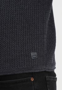 Ciemnoszary sweter o textured powierzchni z wełnianym wzorem, z małą metką z logo na dole i prostym krojem.