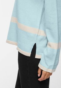 Pull bleu clair à rayures horizontales crème, avec des fentes latérales, des manches longues et une texture douce. Associé à un pantalon noir.