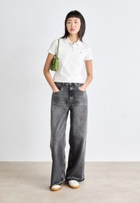 Chemise polo blanche, taille haute, jean noir délavé à la coupe ample avec des ourlets effilochés, associé à un sac à épaule vert et des baskets blanches.