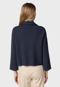 Navy blue cropped jacket with a collar, three-quarter sleeves, and rolled cuffs, made from a textured fabric. Worn over beige trousers.