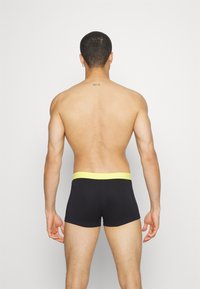 Black boxer briefs with a flat yellow elastic waistband, fitted design, and smooth texture. The back view shows minimal branding detail.