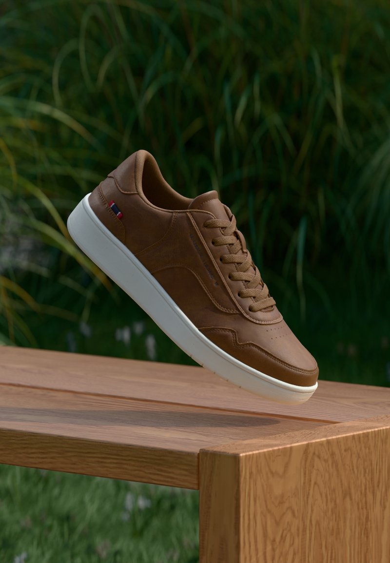 Καφέ δερμάτινο αθλητικό παπούτσι με άσπρη ελαστική σόλα, με στρογγυλά κορδόνια, διάτρητο σχέδιο στη μύτη και ένα μικρό κόκκινο και μπλε διακοσμητικό στη πλευρά.