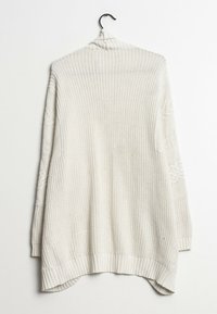 Lys beige strikket cardigan med høj krave, tekstureret mønster, ribbet kant og asymmetrisk snit. Hængende på en sort krog mod en hvid baggrund.