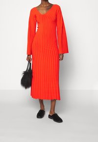 Robe midi rouge côtelée avec manches longues et col en V. Associée à un sac à main en fourrure noire et des chaussures noires à enfiler. Texture lisse.