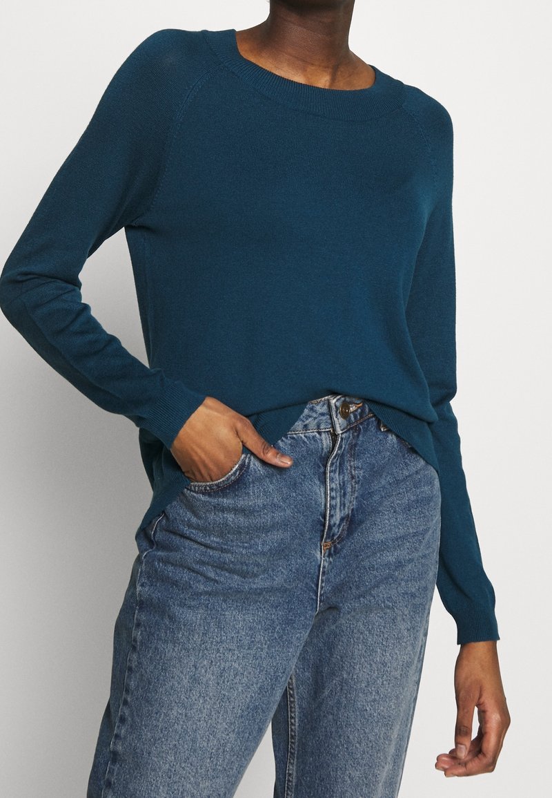 Maglione a maglia teal con scollo rotondo e maniche lunghe, abbinato a jeans blu a vita alta. Il maglione ha una vestibilità rilassata e bordi a coste.