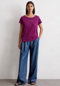 Paarse T-shirt met de tekst "IT'S ON US", gecombineerd met losse blauwe denim broek met een hoge taille en brede pijpen.