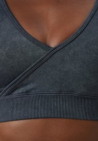 Tumši pelēks sporta krūšturis ar V-veida kakla dizainu, teksturētu audumu un ribotu apakšmali. Materiāls izskatās elastīgs un gluds.