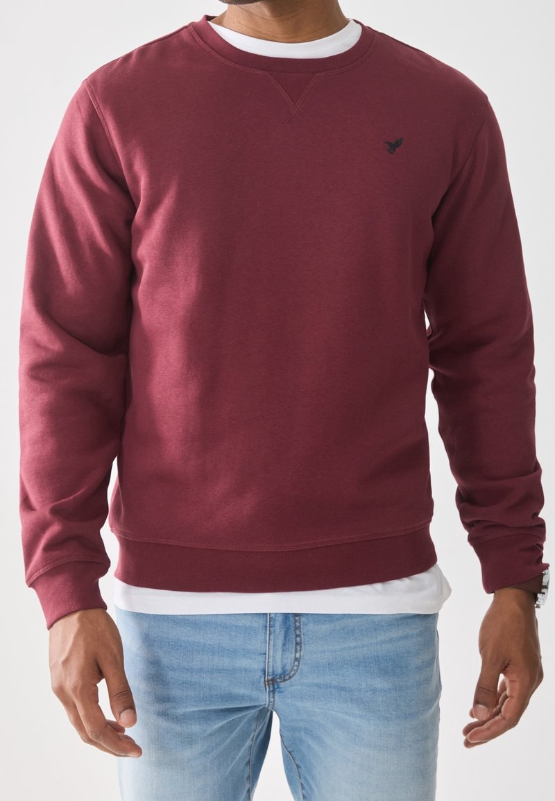 Homme portant un sweat bordeaux sur une chemise blanche, associé à un jean bleu clair, debout les bras détendus le long du corps.