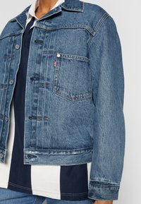 Blå denimjacka med en klassisk design som har spetsig krage, framfickor och knäppning med knappar. Materialet har en strukturerad yta.