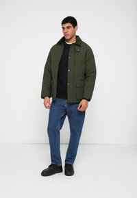 Quilted green jacket with a stand collar, front snap closure, and two pockets, paired with a black sweater and blue jeans.