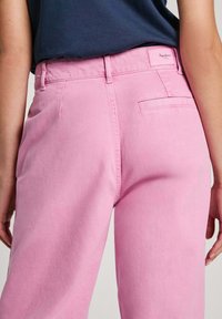 Calças de algodão rosa com um design de perna reta, apresentando um bolso traseiro e um emblema da marca na cintura. Textura suave, ajuste de cintura média.