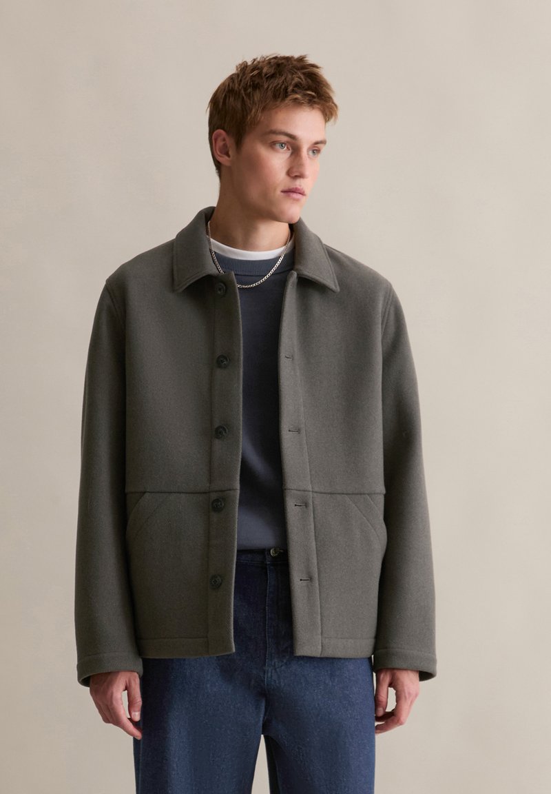 Grijze wollen jas met een kraag, knoopsluiting, twee voorzakken en een strakke, gestructureerde silhouette. G gedragen over een marineblauwe trui.