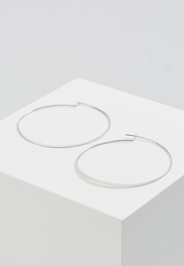 Paire de boucles d’oreilles fines en argent avec fermoir simple posée sur un socle blanc sur fond gris.