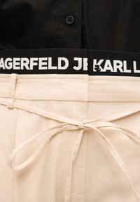 Κοντινό πλάνο μπεζ παντελονιού δεμένου με λεπτή υφασμάτινη ζώνη, που δείχνει τη μαύρη ζώνη μέσης με λευκή επιγραφή "KARL LAGERFELD JEANS" και λογότυπο.