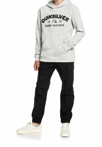 Persona che indossa un felpa grigia chiara Quiksilver con la scritta "Surf Goods", pantaloni cargo neri e sneaker bianche, in piedi con una mano in tasca.