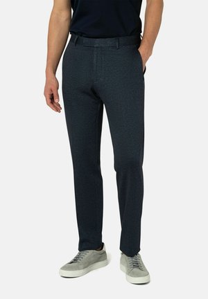 Mann in dunkelblauen Slim-Fit-Hosen und grauen Sneakers, der mit einer Hand in der Tasche vor einem schlichten Hintergrund steht.
