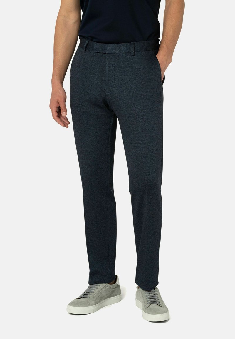 Man in een donkerblauwe slim-fit broek en grijze sneakers, staand met één hand in de zak tegen een effen achtergrond.