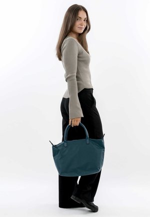 Teal fabric tote bag with two handle straps, structured shape, and a zipper closure. Worn casually with a light gray top and black pants.