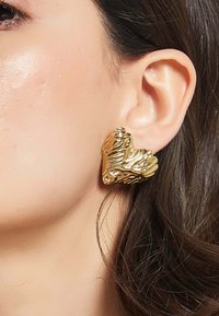 Guldtonade hjärtformade örhängen med detaljerade bladmönster och texturerade ytor, placerade mot örat, som reflekterar ljus.