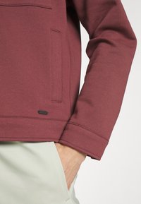 Burgundy tröja med ficka fram, ribbade ärmslut och en liten svart logotagg. Slät tygtextur, matchad med ljusgröna byxor.