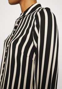 Blusa a righe bianche e nere con colletto e chiusura frontale con bottoni, realizzata in un tessuto testurizzato e con una leggera lucentezza sulle righe.