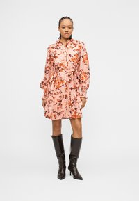 Γυναίκα με πλεγμένα μαλλιά, ντυμένη με ροζ φόρεμα με λουλούδια, μακριά μανίκια και ζώνη, συνδυασμένο με καφέ σκούρα μπότες με τακούνι μέχρι το γόνατο, όρθια.