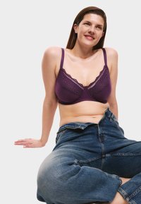 Lila Spitzen-Mikrofaser-Bralette mit verstellbaren Trägern, kombiniert mit hoch taillierten blauen Jeans, präsentiert an einem sitzenden Modell.
