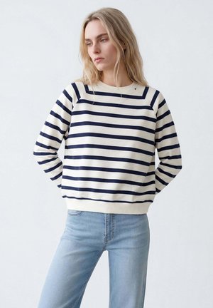 Blonde Frau trägt ein marineblau-weiß gestreiftes Langarmshirt und hellblaue Jeans, steht mit den Händen hinter dem Rücken und schaut nach unten links.