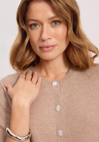 Beżowy sweter z dzianiny z trzema ozdobnymi guzikami w kształcie klejnotów oraz prążkowanym okrągłym dekoltem, uzupełniony srebrną bransoletą na nadgarstku.